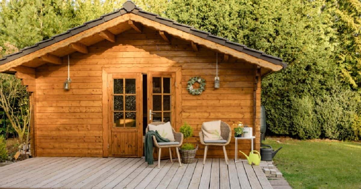 Houten tuinhuis met dubbele deuren op een houten terras, met stoelen en planten ervoor.