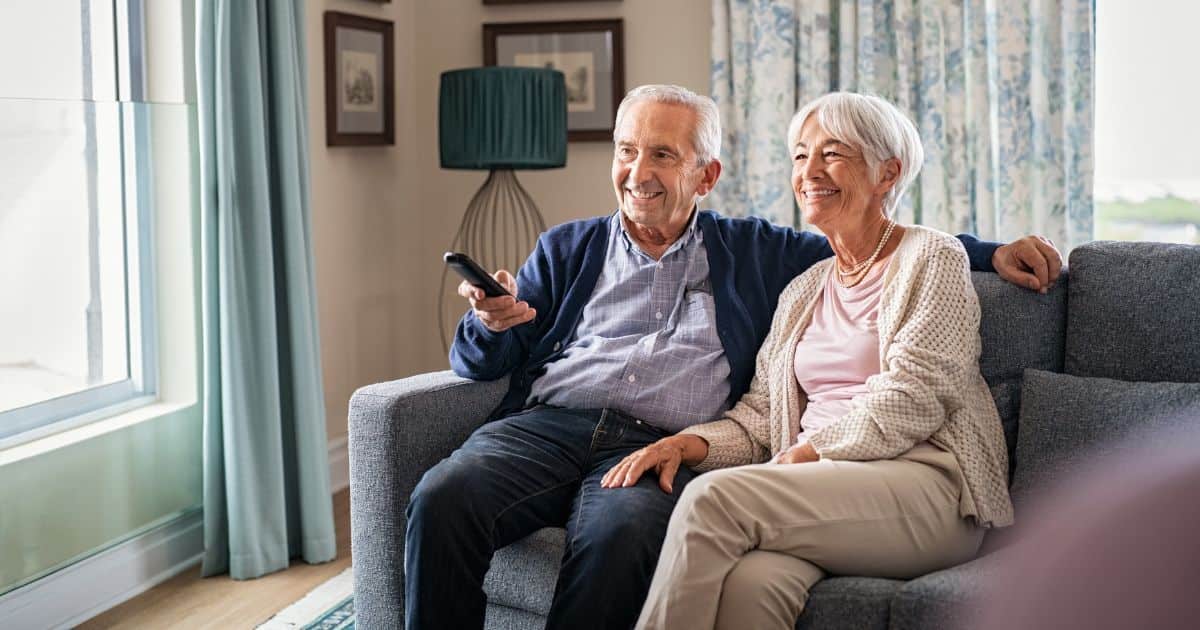 Een oudere dame en een oudere heer zitten samen in een grijze zetel in een woonkamer. De man heeft een afstandsbediening vast. Beide personen glimlachen.