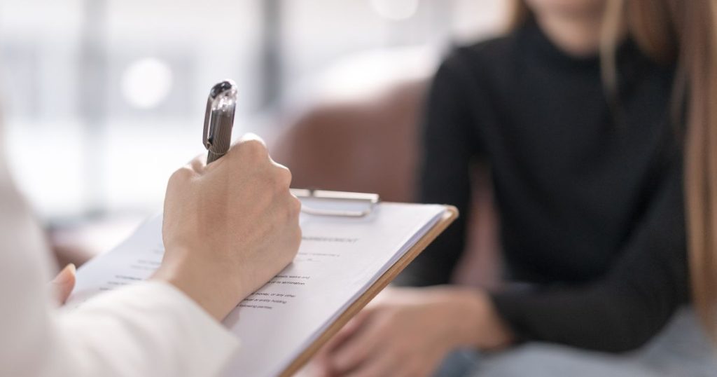 Close-up van het hand van een psycholoog met een pen en een klembord, op de achtergrond een dame met zwarte trui, grijze broek en lang haar.