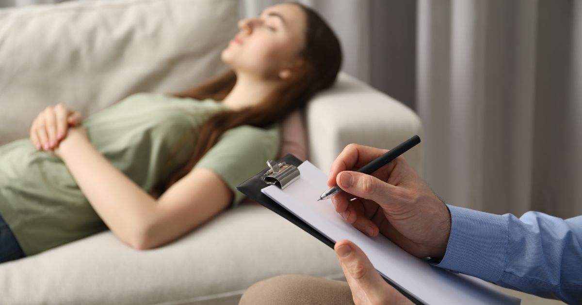 Een dame ligt op een beige sofa. Naast haar zit een man met een blauw hemd en een pen en notitiebord in de handen.