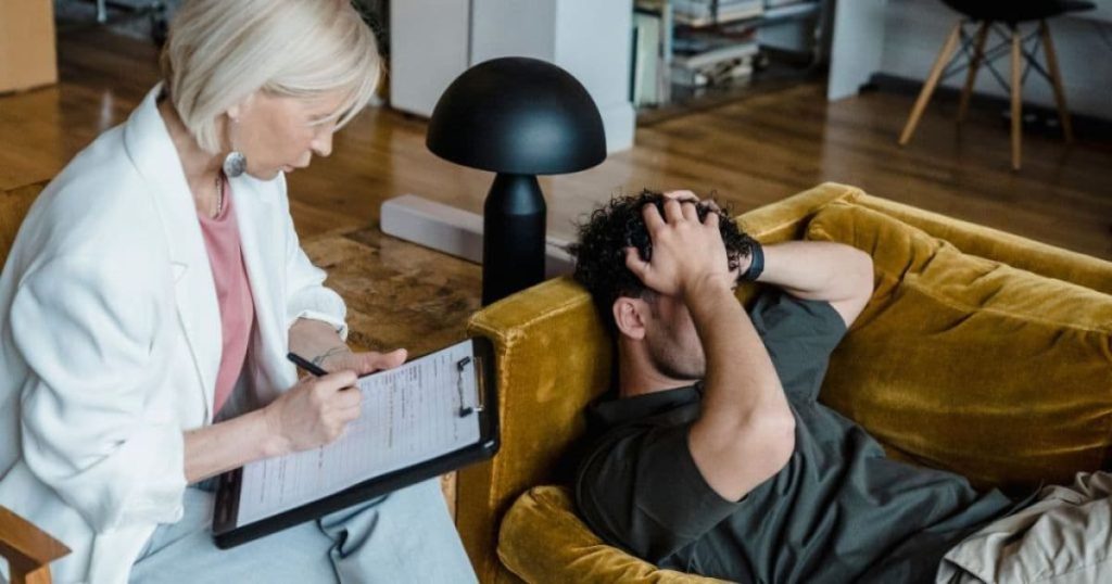 Een man houdt zijn hoofd vast terwijl hij op een sofa languitgerekt ligt. Op een stoel naast hem zit een dame notities te nemen op een blad.