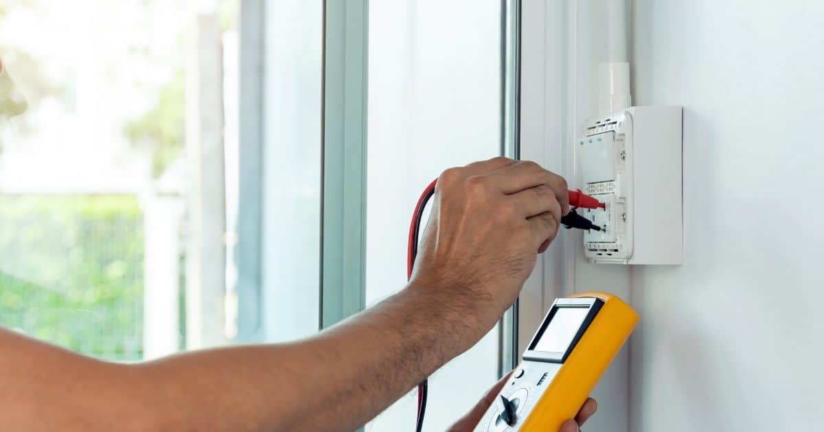 Een persoon voert een testcontrole uit op de schakelaar van een woning. Hij of zij gebruikt daarvoor een stroommeter. Enkel zijn of haar arm is in beeld.