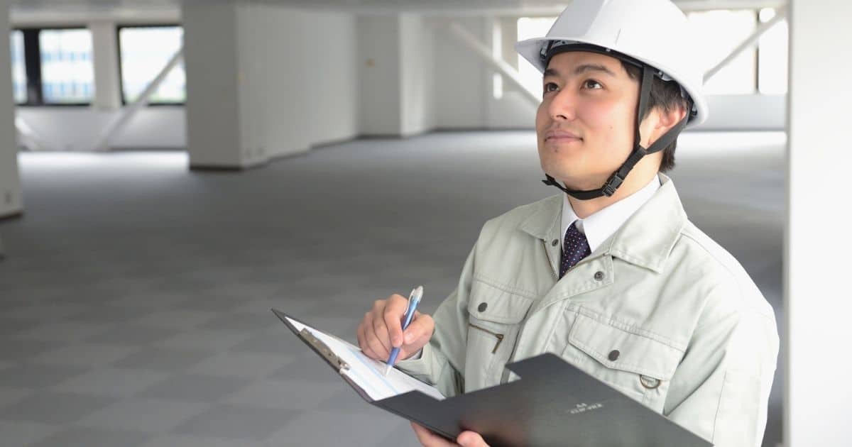 Een technicus in een witte jas en witte veiligheidshelm kijkt omhoog. Hij heeft een zwarte map vast in zijn linkerhand. In zijn rechterhand houdt hij een pen vast. Hij neemt notities.