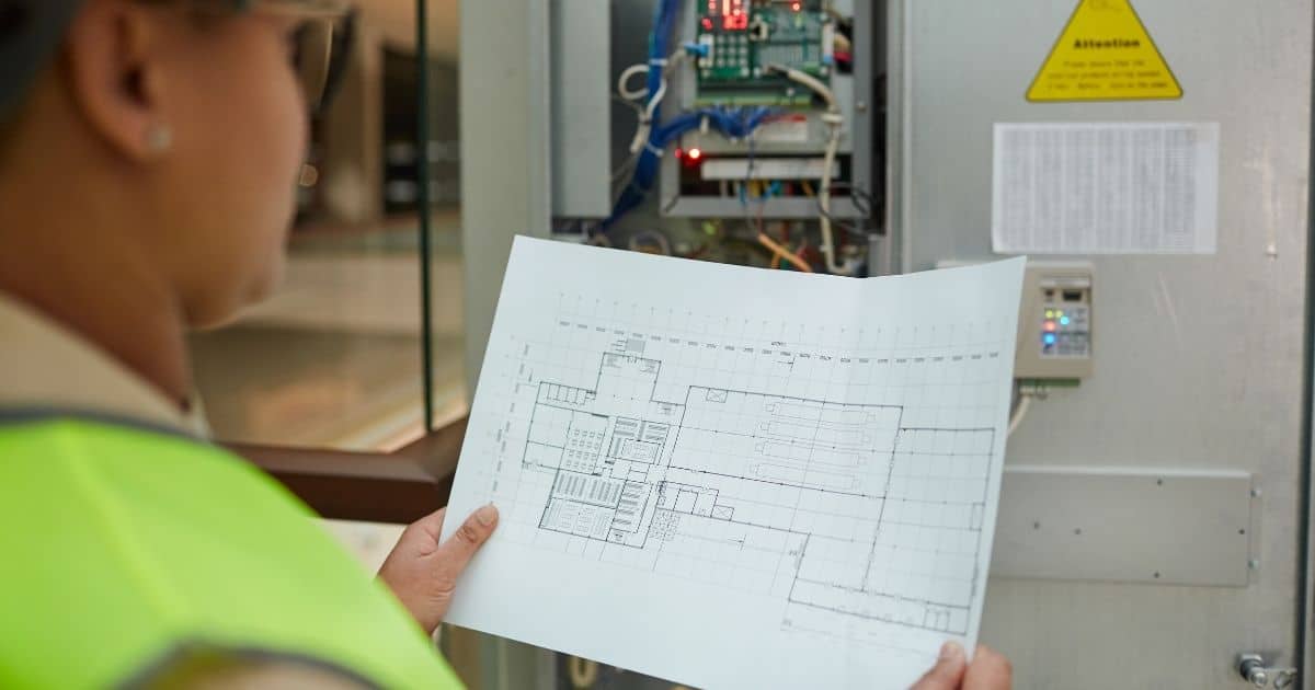 Een elektrisch keurder kijkt na of een meterkast overeenstemt met de situatie op het eendraads- of situatieplan. Ze draagt een fluohesje.