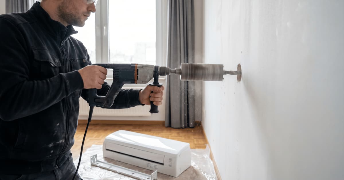 Een technicus met veiligheidsbril boort met een kernboor een ronde doorvoer door een witte binnenmuur, terwijl een airco-binnenunit op een beschermfolie op de grond klaarligt voor montage.