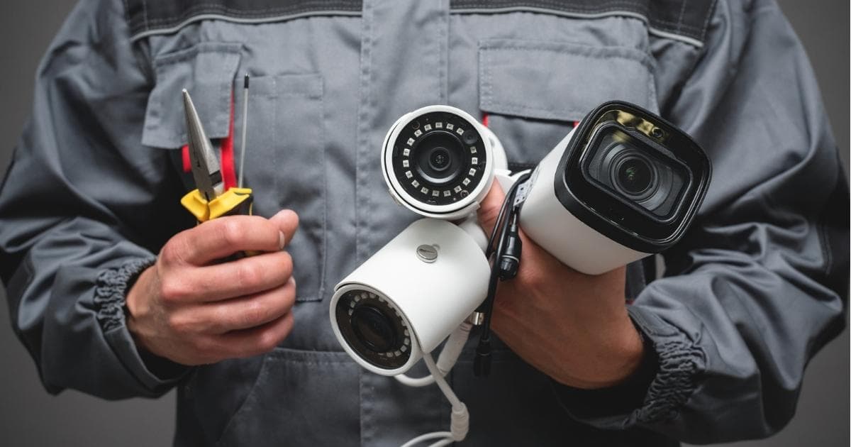Een installateur van camerabewaking in een grijze werkoutfit. In zijn rechterhand heeft hij 3 verschillende soorten bewakingscamera's vast, inclusief de bekabeling. In zijn linkerhand heeft hij gereedschap vast.