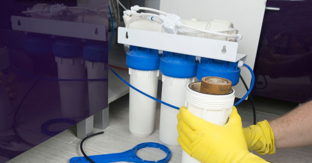 Een installateur met gele handschoenen houdt één van de kokers van een waterfilter vast. De kokers zijn in het wit en hebben een blauwe dop.