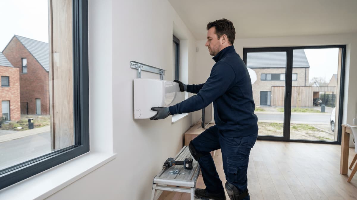 Een technicus in donkerblauw werkpak staat op een trapje en plaatst een witte airco-binnenunit op een metalen muurbeugel in een moderne woning, met door het raam een Belgische nieuwbouwwijk zichtbaar onder bewolkte hemel.