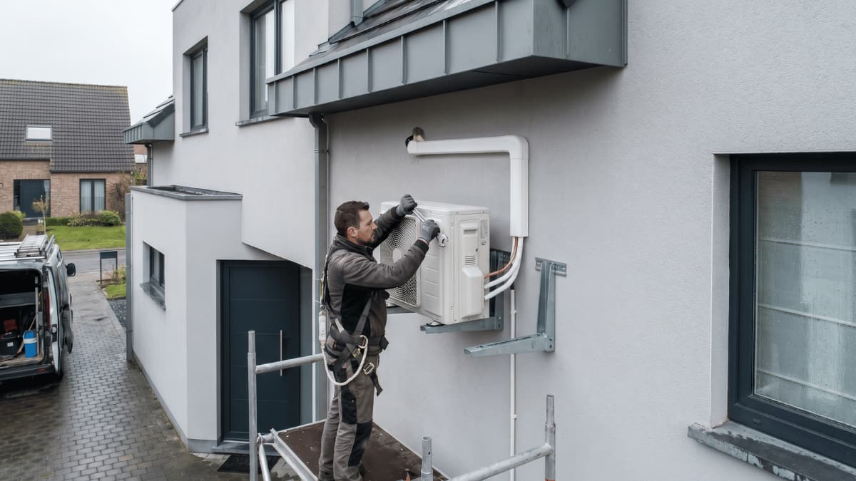 Een technicus in werkkleding en veiligheidsharnas staat op een steiger en bevestigt een airco-buitenunit op metalen muurbeugels aan de lichtgrijze gepleisterde gevel van een moderne woning, met witte leidingkokers en koperen koelleidingen zichtbaar naast het toestel.