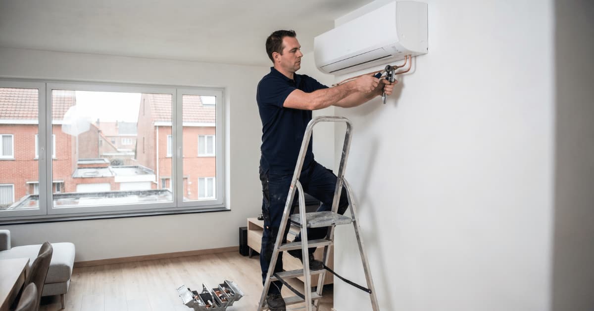  airco-installateur in donkerblauw poloshirt staat op een aluminium trapladder en sluit met een flaretang koperen leidingen aan op een witte wandairco hoog aan de muur in een lichte woonkamer met zicht op Vlaamse bakstenen daken door het raam.