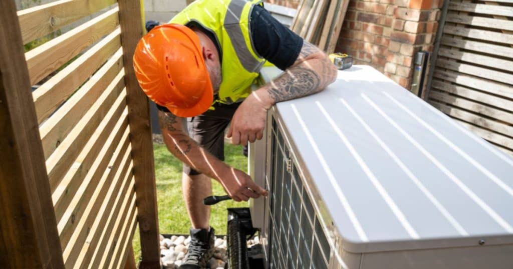 Monteur met oranje helm en fluohesje installeert buitenunit van een warmtepomp naast houten schutting