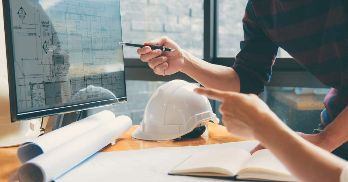 Een interieurarchitect zit samen met een aannemer achter een computer. Op het scherm is een plattegrond zichtbaar. Op het bureau liggen een veiligheidshelm, notitieblok en 2 opgerolde bouwplannen.