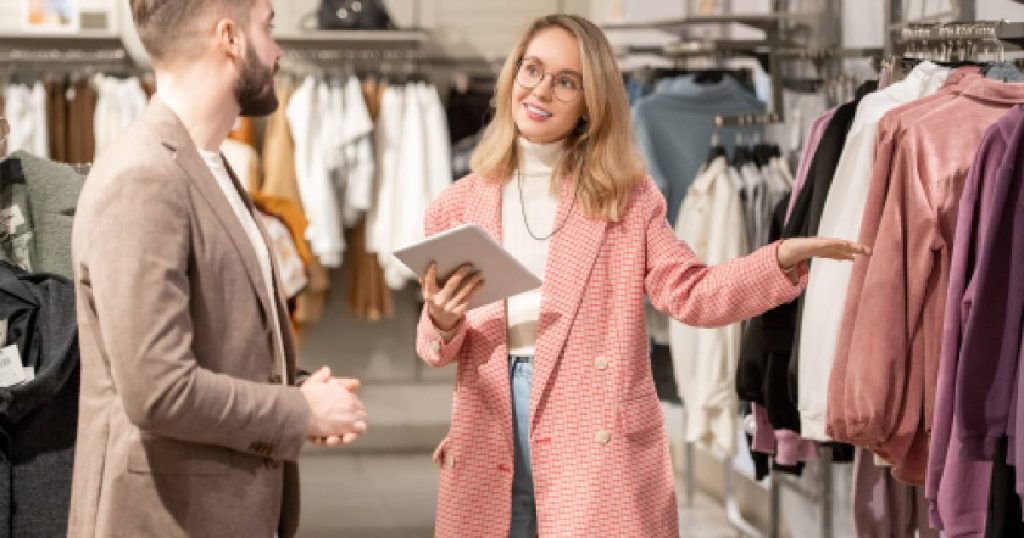 Een blonde verkoopster in een lichtroze jas houdt een iPad kassa vast en toont een klant een roze trui aan een kledingrek in een kledingwinkel. 