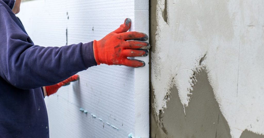 Een gevelspecialist brengt dikke harde isolatieplaten aan tegen een gevel. Hij draagt een paarsblauwe trui en rode werkhandschoenen, bedekt met mortel.