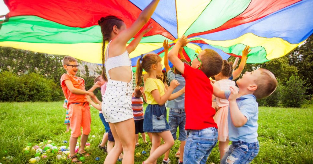 Een groep kinderen van verschillende leeftijden onder een zeil met regenboogkleuren op een grasveld tijdens een activiteit van de jeugdbeweging.