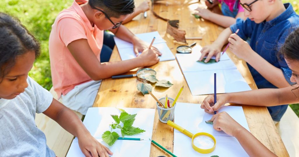 Een groep kinderen in open lucht aan een houten tafel. Ze traceren de omtrek van bladeren tijdens een activiteit van de jeugdbeweging.