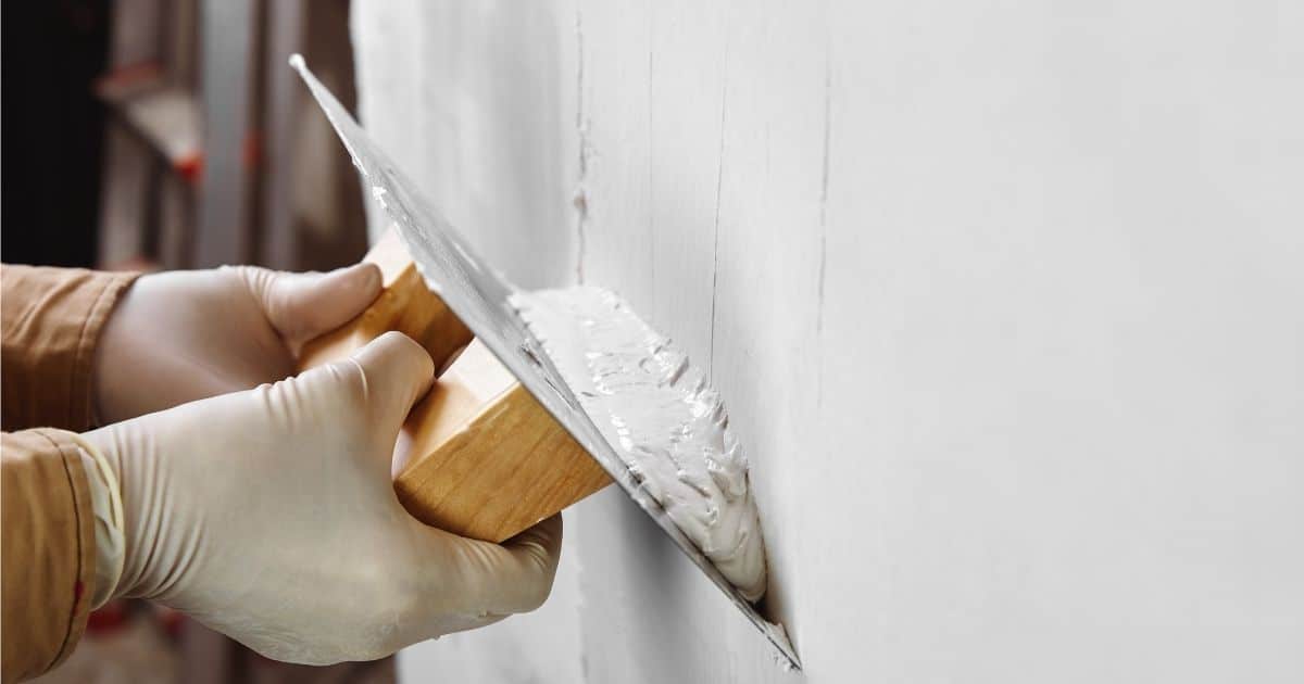 Een stukadoor of plakker brengt pleister aan op een binnenmuur in een nieuwbouwwoning. Hij of zij gebruikt een spaan om het materiaal gelijkmatig aan te brengen. De stukadoor draagt latex handschoenen.