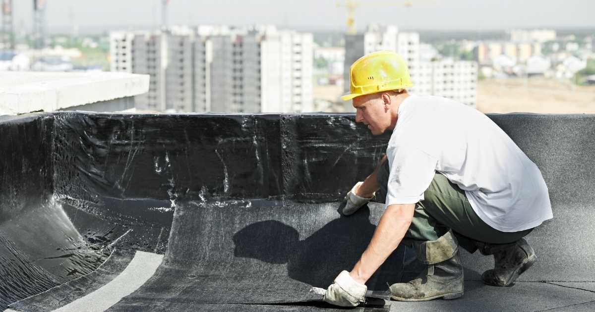 Een dakspecialist in wit t-shirt, groene werkbroek, laarzen en gele veiligheidshelm brengt een strook dakleer aan op een plat dak.