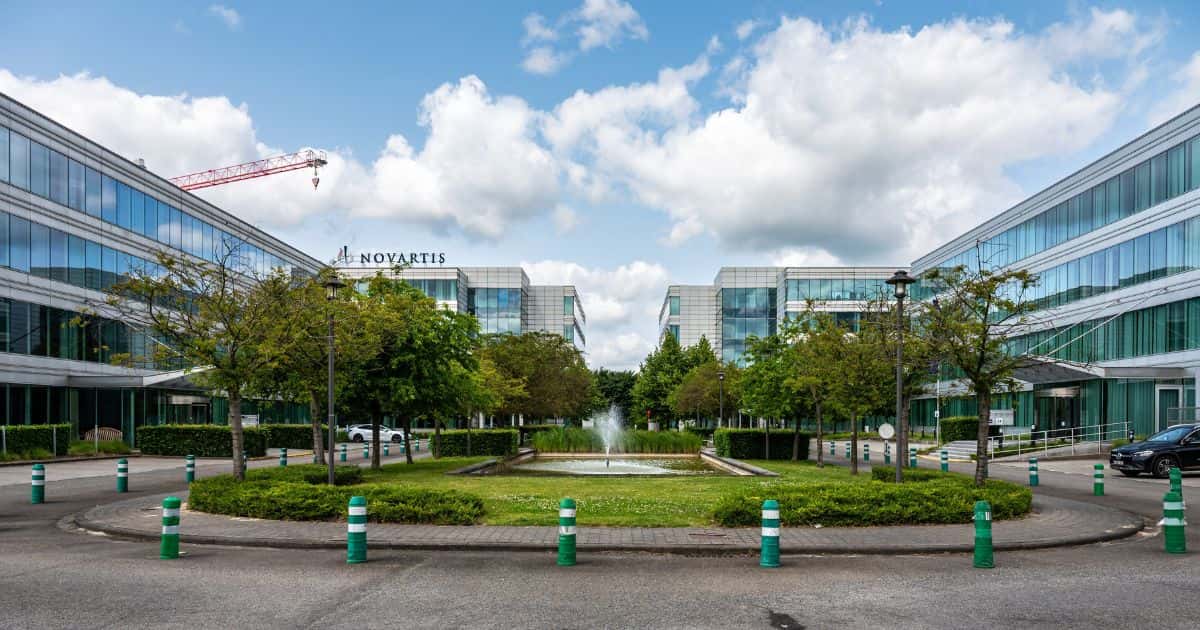 Aan weerszijden van het centrale parkje met waterfontein staan glazen kantoorgebouwen. Op de achtergrond is het logo Novartis zichtbaar, en ook een oranje bouwkraan.