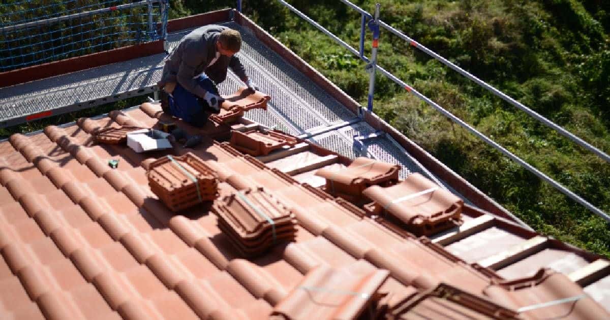 Een dakwerker legt vlakke keramische dakpannen op het dak van een woning. Hij gebruikt een steiger om het dak veilig te bereiken.