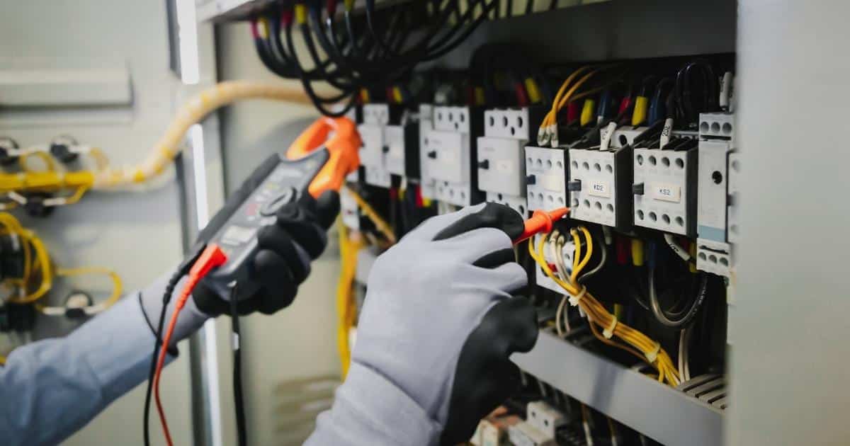 De twee handen van een AREI-keurder met grijs-met-zwarte handschoenen voor een meterkast. In de linkerhand een zwart-met-oranje spanningsmeter, in de rechterhand een oranje plug.