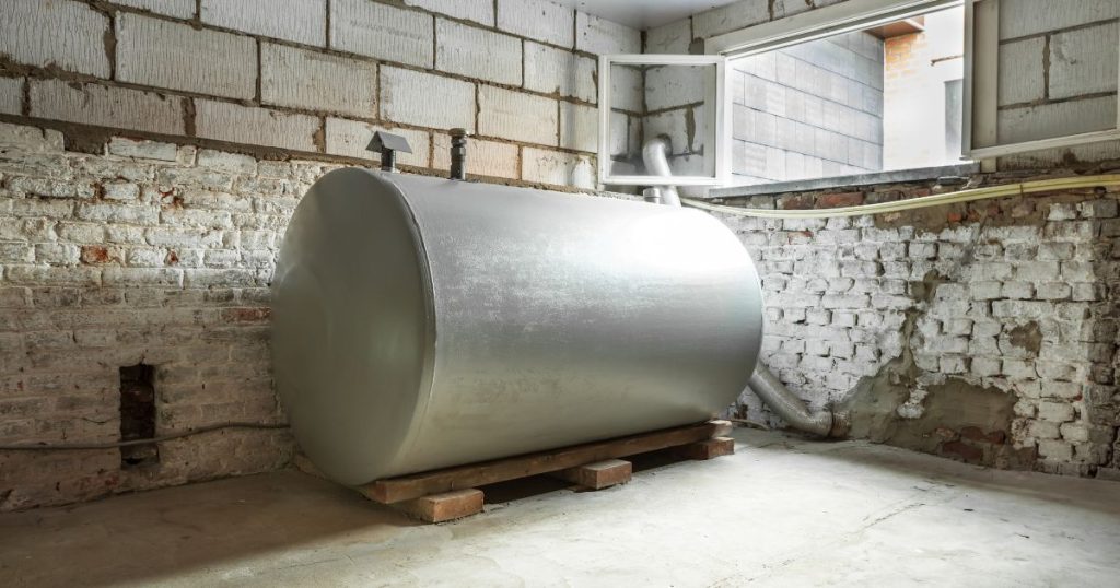 Een grijze, metalen stookolietank of mazouttank in een verluchte kelder met witte bakstenen muur en betonnen vloer.