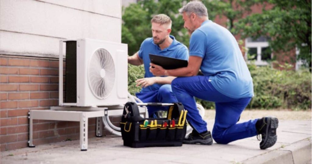 2 mannen in blauwe werkkledij bekijken de buitenunit van een airco: 1 iemand sleutelt aan de unit, de andere houdt een zwart klembord vast. Op de grond staat een tas met werkgerief.