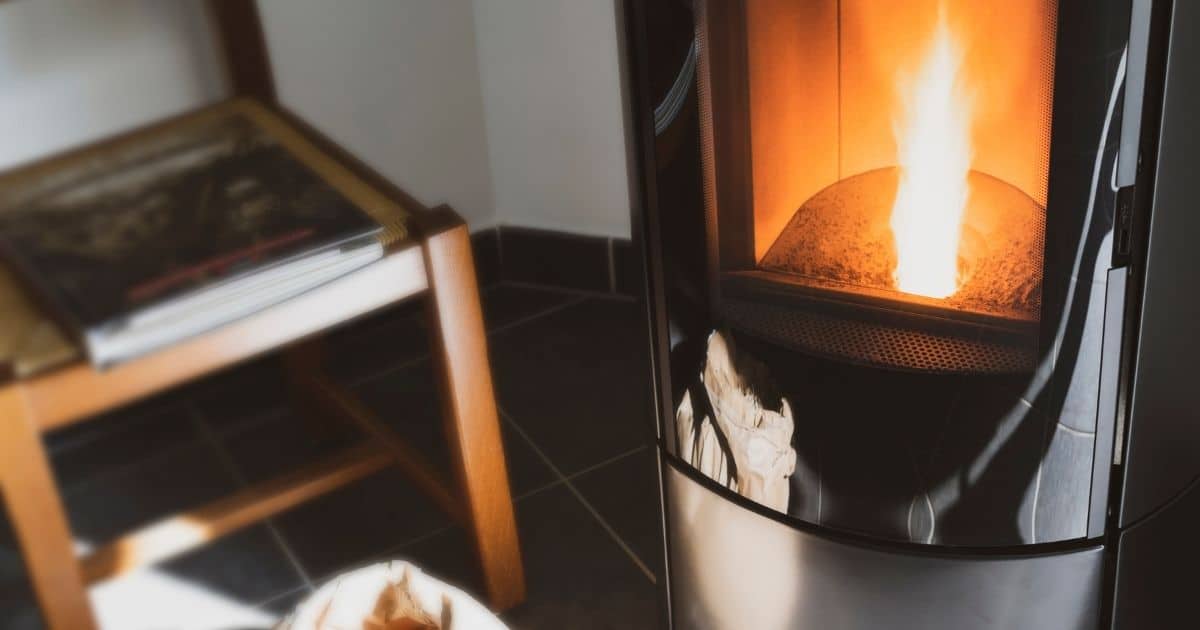 Een ijzeren pelletkachel met brandende pellets op een vloer met donkere tegels. Naast de kachel staat een stoel waarop een boek ligt.