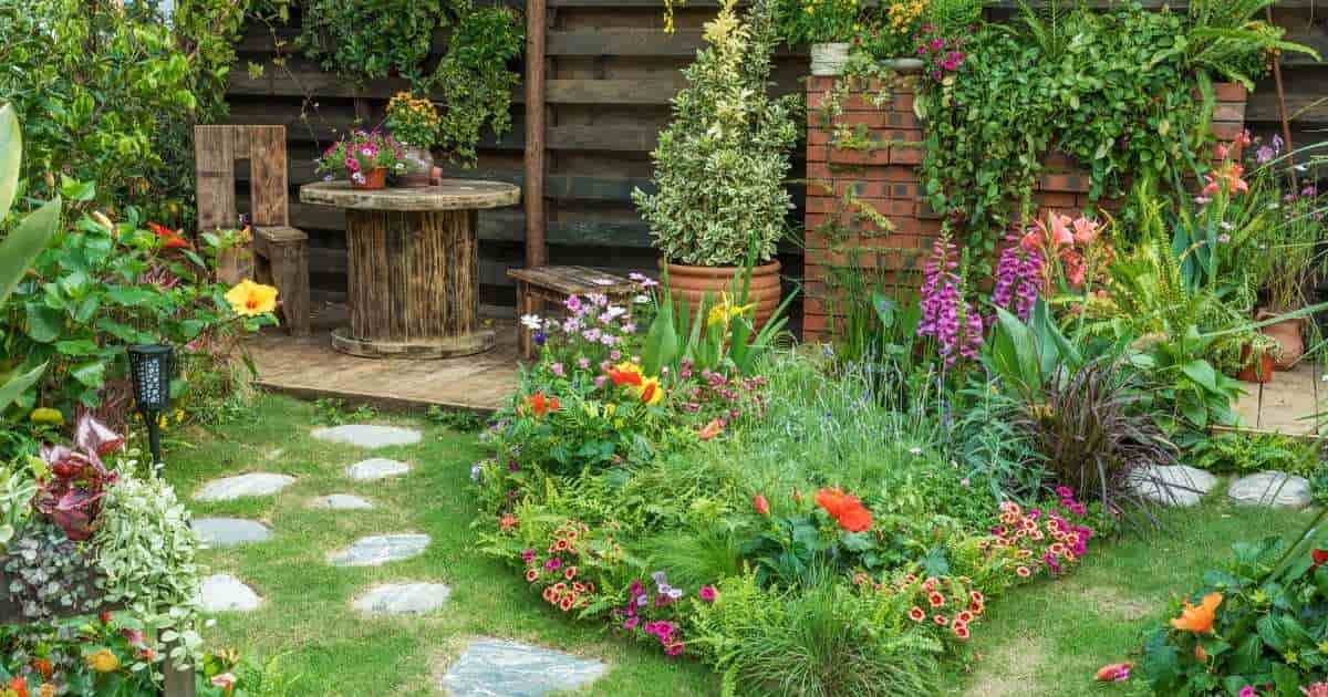 Een kleine maar weelderige tuin met bloemenperken en stapstenen. Achterin de tuin is een kleine zithoek aangelegd met houten terrasvloer, een tafel, stoeltje en zitbankje.