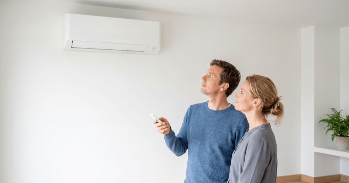 Een man in een blauwe trui richt een witte afstandsbediening op een wandgemonteerde airco-binnenunit terwijl een vrouw naast hem meekijkt in een lichte woonkamer met een groene plant op de achtergrond.