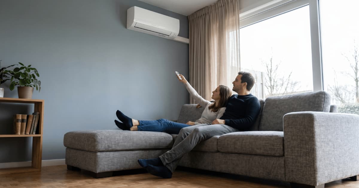Een koppel zit ontspannen op een grijze hoekbank terwijl de vrouw een afstandsbediening richt op de witte airco-binnenunit hoog op de grijsblauwe muur in een lichte woonkamer met houten vloer.