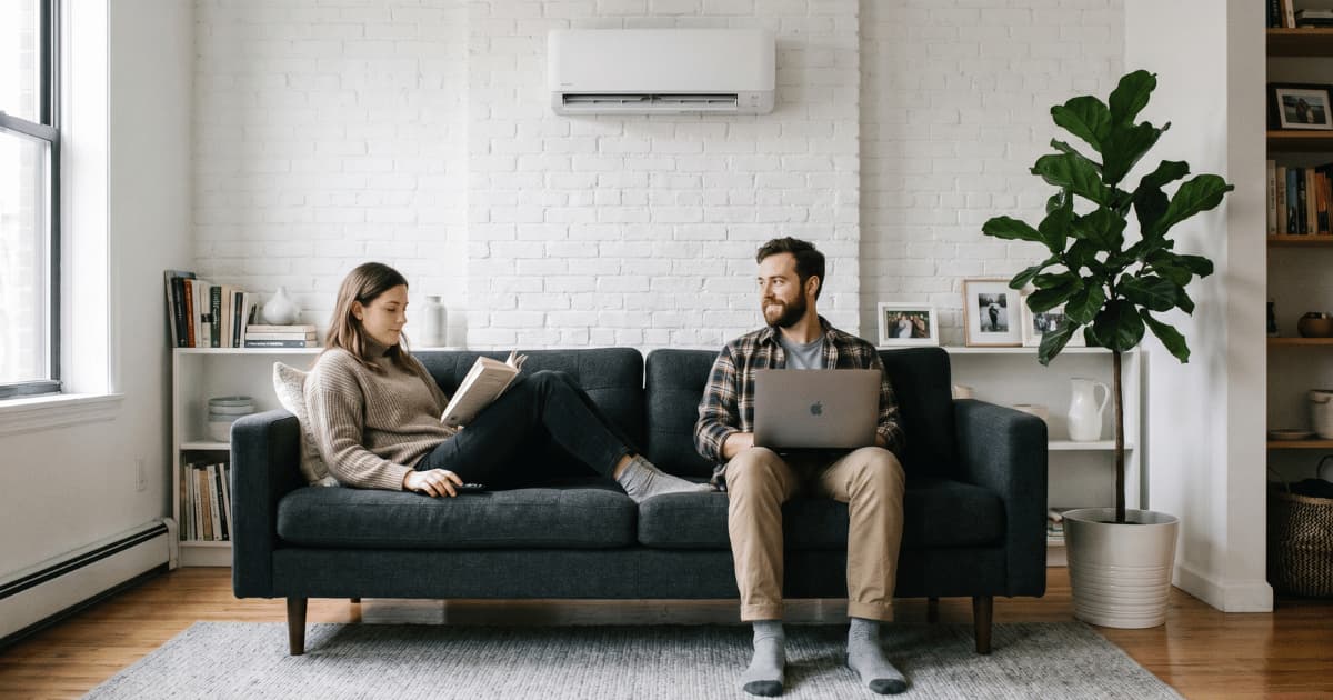 Een vrouw leest een boek en een man werkt op een laptop op een donkergrijze sofa in een gezellige woonkamer met een witte airco binnenunit aan een witgeschilderde bakstenen muur en een grote groene plant ernaast.