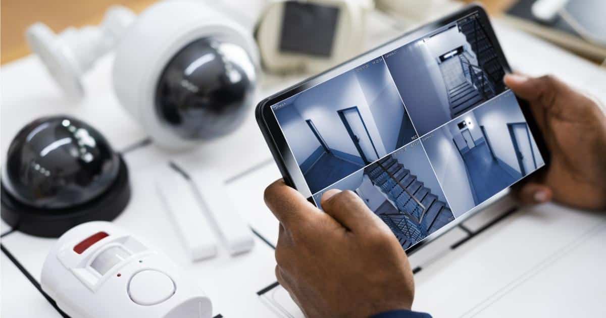 Twee handen houden een tablet vast waarop camerabeelden van verschillende ruimtes in een gebouw te zien zijn. Daaronder een tafel met een kaart van het beveiligingssysteem en verschillende onderdelen van de alarminstallatie: camera's, bewegingssensoren en meer.