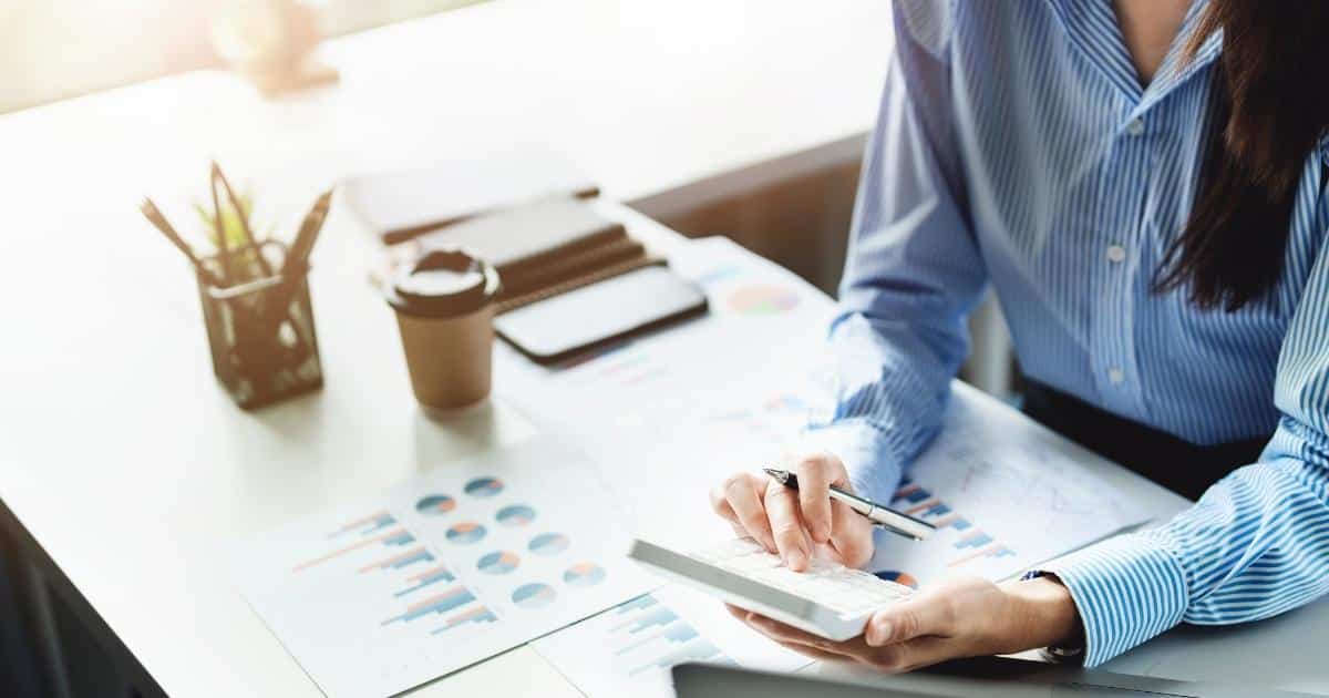 Een zakelijk geklede vrouw met bruin haar in een verticaal, wit met blauw gestreept hemd maakt berekeningen met een rekenmachine. Voor haar op tafel liggen documenten met grafieken. Op de witte tafel staan ook een houder met schrijfgerei, een wegwerp kop koffie, een smartphone en een stapel notitieboekjes.