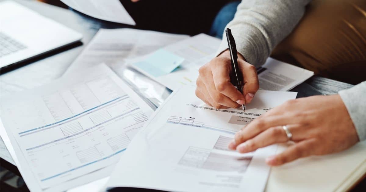 Twee handen boven een stapel documenten. In de rechterhand heeft de persoon een zwarte pen vast. Aan de linkerhand zit een zilveren ring.