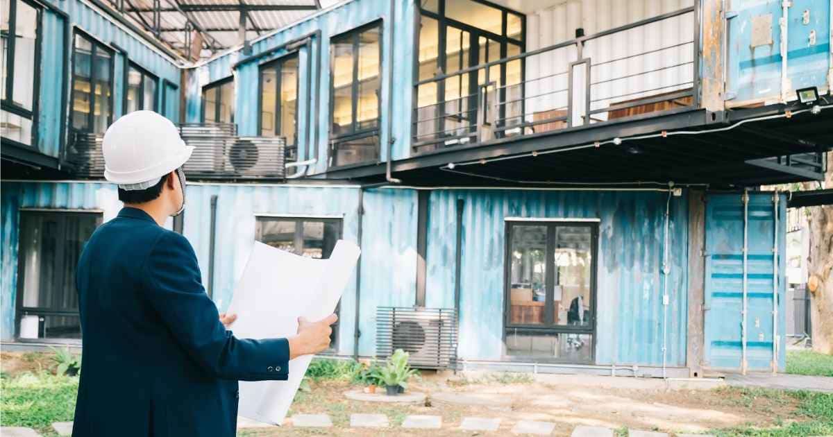 Een architect met bruin haar onder een witte veiligheidshelm en een blauw kostuumvest houdt een groot bouwplan vast. Hij kijkt naar een grote, oude woning met 2 verdiepingen, gebouwd uit blauwe metalen containers. 