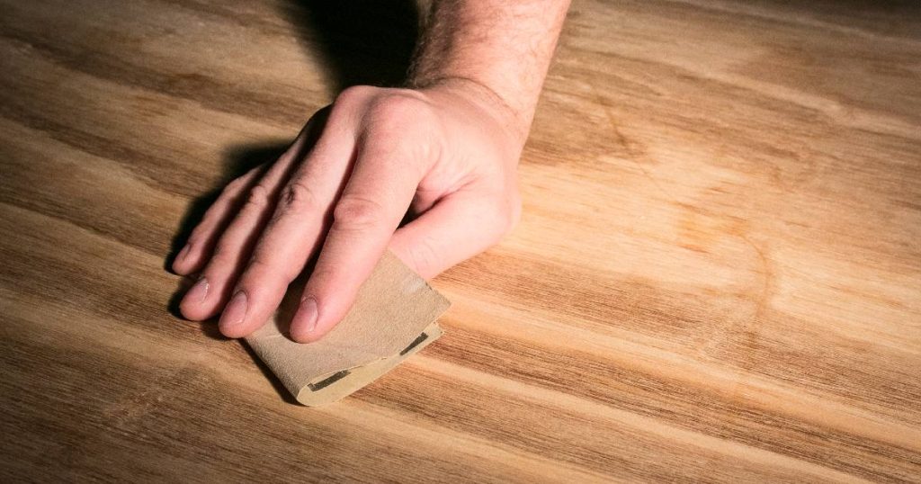 Beeld van de hand van een man op een houten vloer. De man gebruikt een stukje schuurpapier om krassen op een parket te verwijderen.