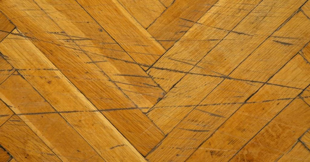 Bovenaanzicht van een gele houten vloer met visgraatpatroon en verschillende diepe krassen.