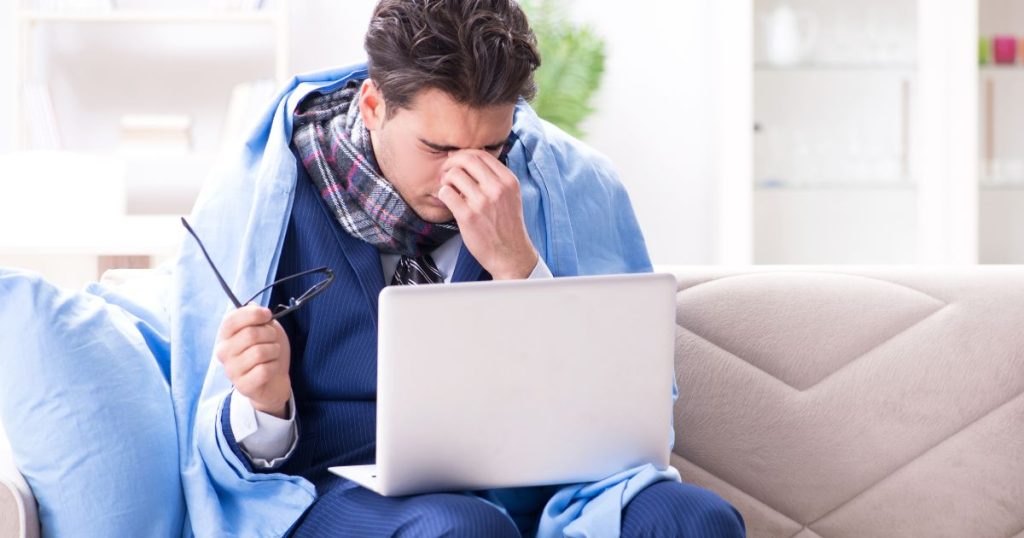 Een zieke werkgever zit op een witte zetel met een laptop op de schoot. Hij draagt een blauw vest, een geruite sjaal, en een blauwe broek. Op de zetel ligt een blauw hoofdkussen, en rond zijn schouders een blauw deken. Hij heeft zijn bril vast in zijn rechterhand. Zijn linkerhand houdt hij tegen zijn hoofd terwijl hij fronst. 