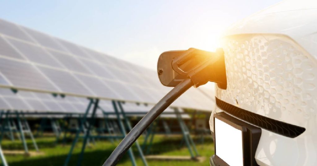 De achterkant van een elektrische bedrijfswagen met ingeplugde laadpaal, op de achtergrond zonnepanelen die het bedrijf voorzien van groene energie voor de laadpalen.