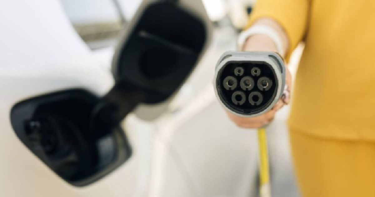 Het kopstuk van een laadkabel (1-fase of 3-fase) in de hand van een dame in gele jurk naast een witte elektrische auto.