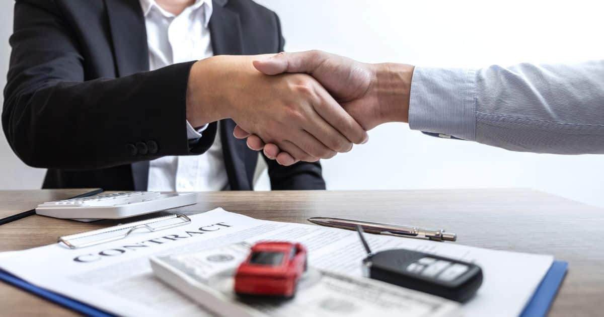 Een ondernemer schudt de hand van een vertegenwoordiger van een leasebedrijf. Onder hun handen, op een houten tafel, ligt een klembord met het leasecontract, een rekenmachine, een pen, autosleutels en een rode miniatuurwagen.