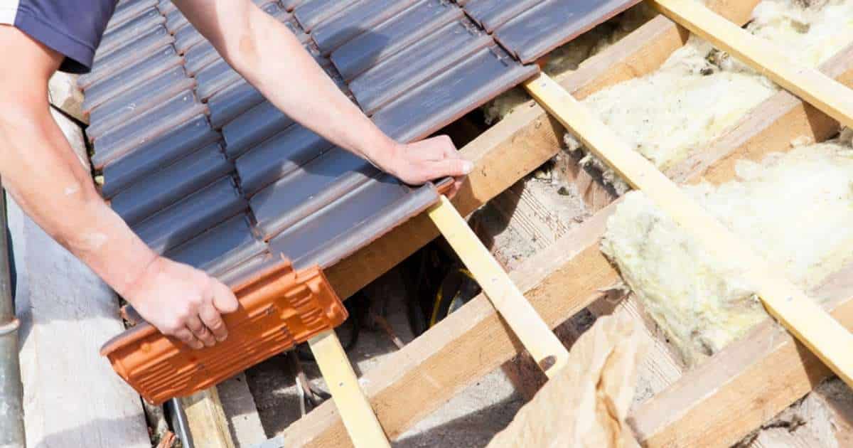 Een dakwerker legt of haalt zwarte dakpannen van het dak. Onderliggend bevindt zich een houten structuur met tussen het dakgebinte stroken glaswol of steenwol.