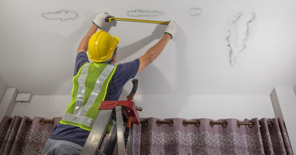 Specialist op ladder bezig met lekkage opsporen en vochtschade meten aan plafond met meetlint.