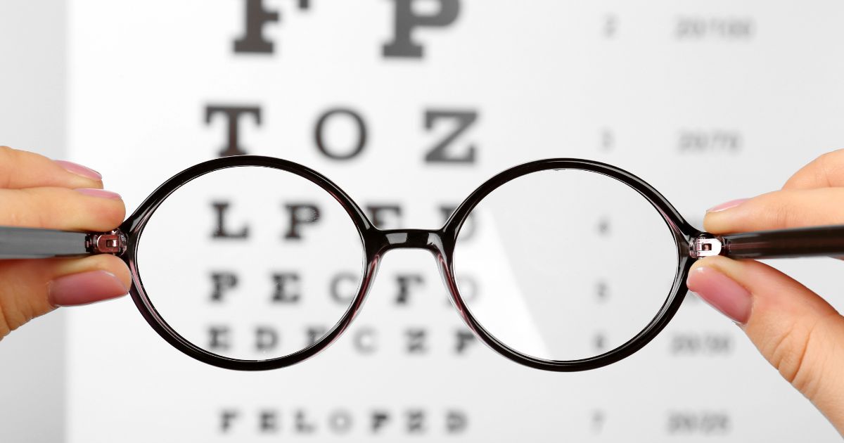 Twee handen houden een bril met zwart montuur voor een leestest: een blad met letters in verschillende groottes. De letters zijn wazig.