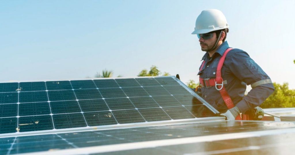 Een installateur met veiligheidsriem en witte helm legt zonnepanelen op een plat dak.