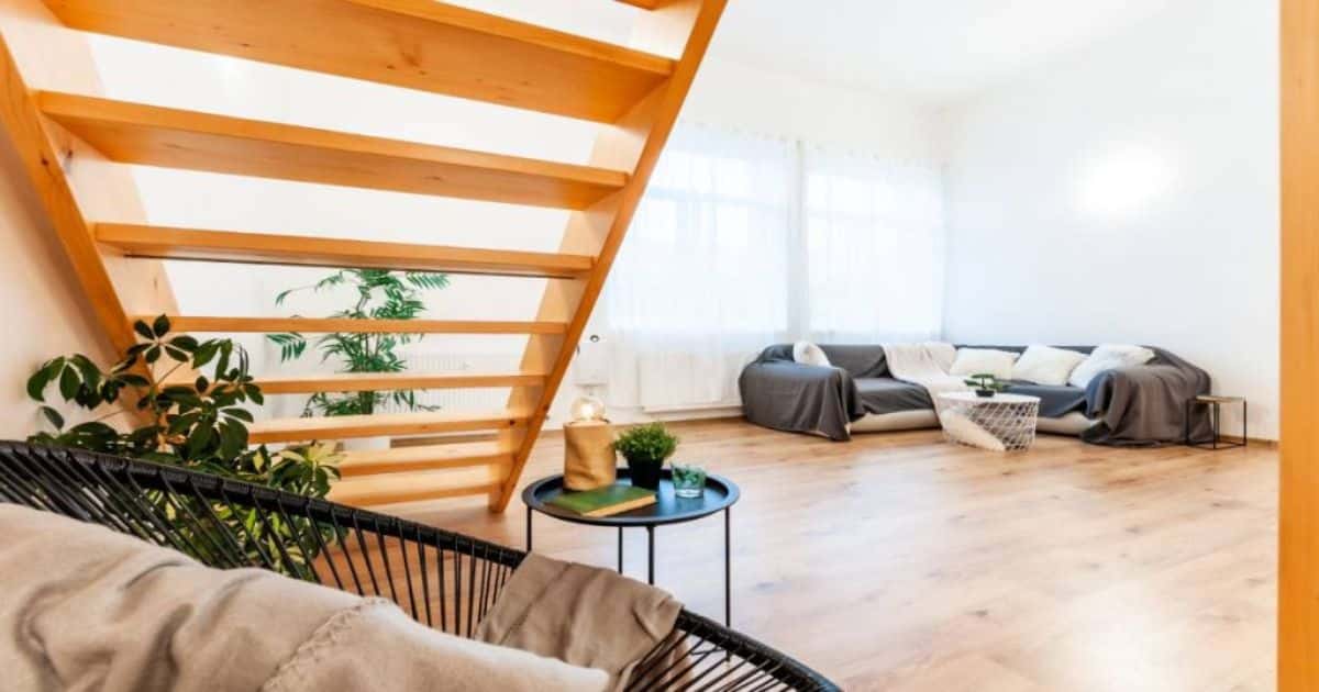 Een lichte houten ruimtebesparende steektrap in een witte woonkamer met zetels en een salontafel. Onder de trap staat een plant, een mini salontafel en een speciale zitstoel.