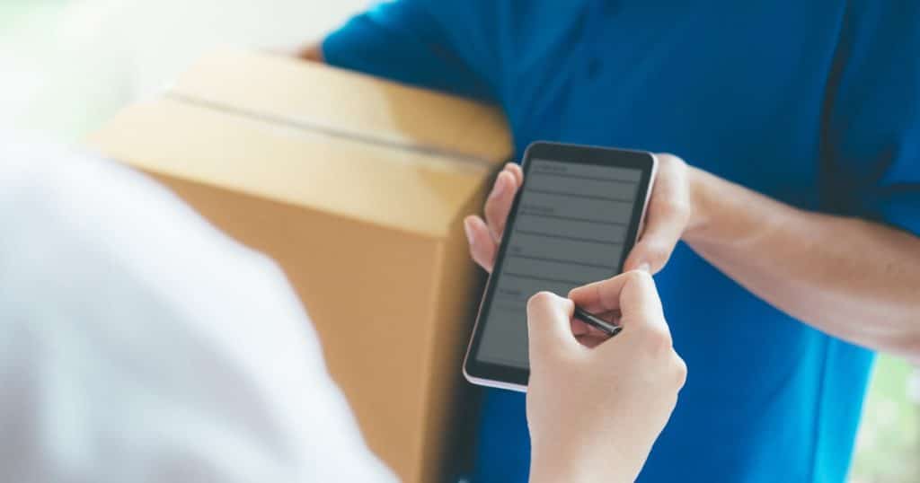 Een man met een blauw hemd gebruikt zijn smartphone om een klant te laten bevestigen dat hij een pakketje heeft ontvangen. De smartphone is gelinkt aan het uitgebreide POS-systeem van een Lightspeed kassa.