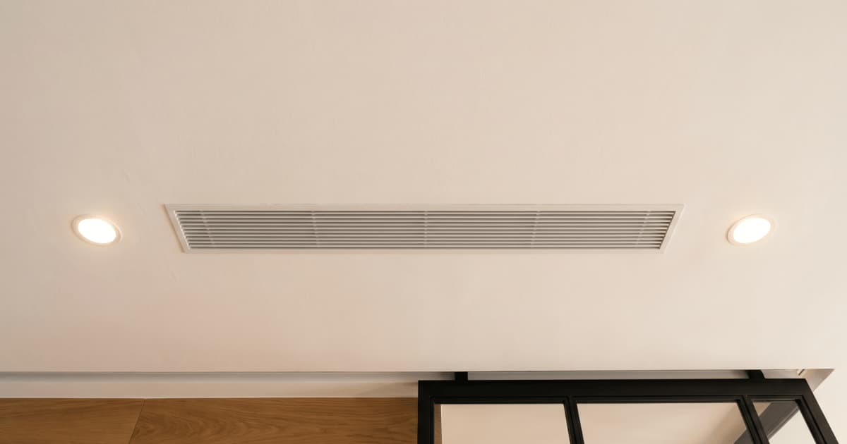 Een lang rechthoekig ventilatierooster met fijne horizontale lamellen zit vlak ingebouwd in een wit plafond met aan weerszijden een ronde inbouwspot terwijl onderaan houten wandpanelen en een zwart stalen glaspartitie zichtbaar zijn.