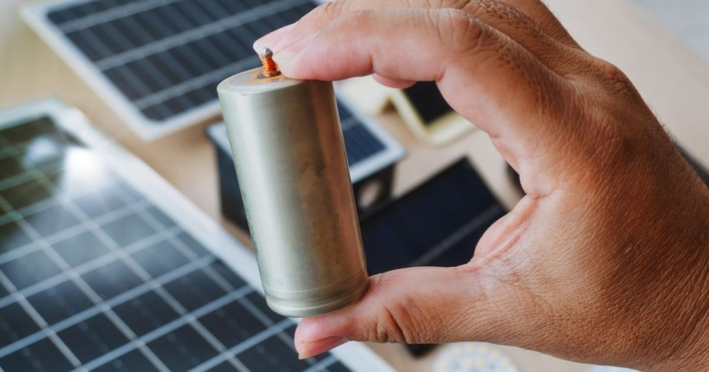 Een hand houdt een kleine batterij met ijzerfosfaat boven een aantal kleine zonnepanelen.